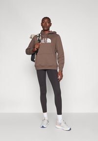 Sweatshirt com capuz castanho e logótipo branco, leggings pretas e sapatilhas cinzentas. Modelo carrega uma mochila escura e está de pé contra um fundo simples.