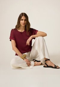 Top corto burdeos con detalles en los hombros, combinado con pantalones anchos blancos y sandalias negras. Pulsera dorada en la muñeca, texturas suaves.