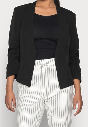 Kvinde iført en sort blazer med rynkede ærmer over en sort top, kombineret med hvide snørede bukser med lodrette sorte striber.