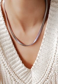 Collana argento con un design liscio e arrotondato, appoggiata su un maglione lavorato a maglia di colore chiaro, che mette in risalto una finitura metallica luccicante.