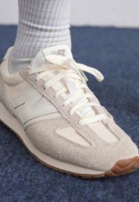 Nærbilde av en lysfarget sneaker med teksturert overdel, som har en fremtredende «N»-logo, som ligger på et blått teppe.