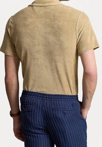 Man som bär en ljusbrun mockakortaärmad skjorta och blå randiga byxor med händerna i fickorna, sedd bakifrån.