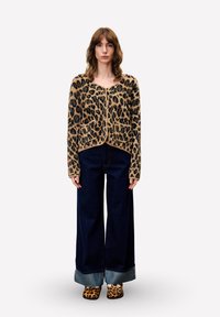 Vrouw die staat in een luipaardprint cardigan, met wijde donkere spijkerbroeken met omslagen en bijpassende luipaardprint schoenen op een effen achtergrond.