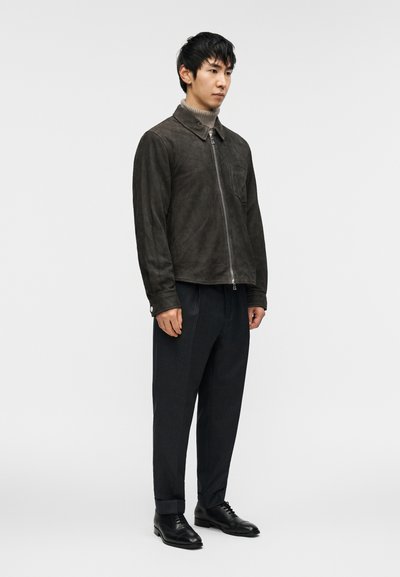 Jachetă maro din piele întoarsă cu fermoar frontal, buzunar lateral și guler, asortată cu pantaloni în dungi subțiri închis la culoare și pantofi negri din piele.