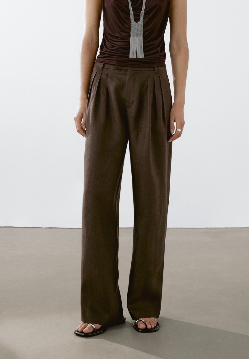 Personne portant un pantalon large marron foncé, un haut noir sans manches, un long collier gris, une bague argentée, et des sandales noires à bouts ouverts, debout sur un sol gris.