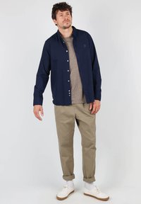 Chemise bleue à boutons, portée par-dessus un pull col rond beige clair, avec un pantalon kaki et des baskets blanches. Présente un col classique et des manches retroussées.