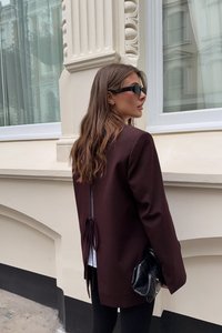 Blazer bordeaux con dettaglio legato, abbinato a leggings neri e una piccola borsa nera. Il modello indossa occhiali da sole neri eleganti.