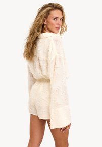 Femme aux longs cheveux blonds ondulés portant une combinaison blanche à manches longues texturée avec de larges manches, se retournant sur son épaule devant un arrière-plan blanc.