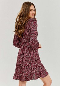 Kvetinové zavinovacie šaty s dlhými rukávmi, zúženým pásom a splývavou sukňou v tmavo ružovej farbe s malými bielymi a zelenými kvetinami.