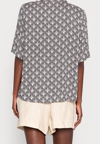 Koszula z krótkim rękawem w czarno-białe wzory z geometrycznym motywem, w połączeniu z lekkimi beżowymi szortami, prezentowana z tyłu.