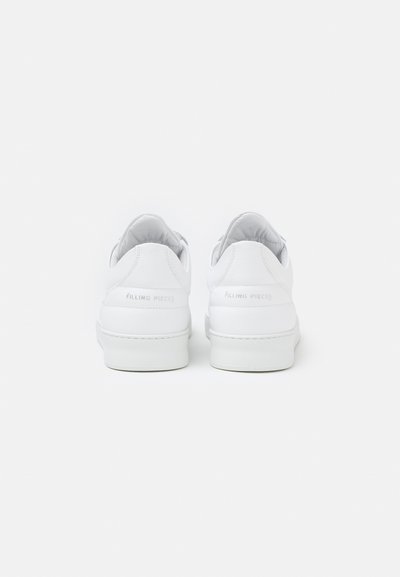 Zapatos deportivos de piel blanca con textura suave, punta redondeada y diseño minimalista. La vista trasera muestra el logo en relieve en el talón y un collar acolchado.