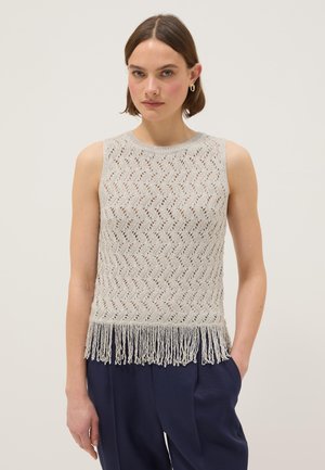 Mujer con un top sin mangas de punto gris claro con patrón en zigzag y dobladillo con flecos, combinada con pantalones azul oscuro contra un fondo liso.