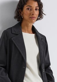 Dark gray tailored coat with a notched collar, single button closure, and side pockets, worn over a light cream textured sweater.
