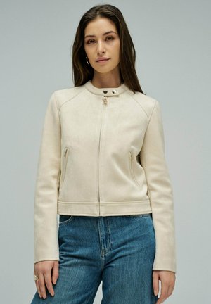 Femme aux longs cheveux bruns portant une veste zippée beige clair avec col pressionné et un jean bleu, debout devant un fond uni.