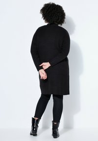Personne aux cheveux bouclés portant un long cardigan noir, un pantalon noir et des bottines noires, debout de dos, les mains croisées derrière le dos.