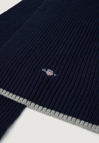 Scarf a coste nel colore blu navy con un accento grigio chiaro alle estremità. Presenta un logo ricamato a colori su un lato. Texture morbida, forma rettangolare.