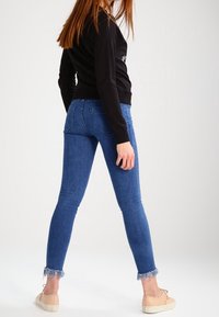 Pantalones vaqueros ajustados azul claro con dobladillos deshilachados, lavado medio y bolsillos traseros, combinados con una camiseta negra de manga larga y zapatos planos beige.