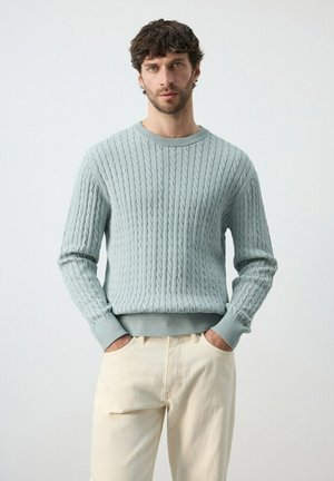 Uomo che indossa un maglione azzurro a trecce e pantaloni color crema con le mani nelle tasche, in piedi davanti a uno sfondo chiaro e semplice.