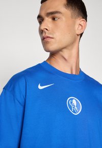 Blå bomulds T-shirt med korte ærmer, der viser et hvidt Nike-logo på skulderen og et rundt hvidt klubemblem på brystet.
