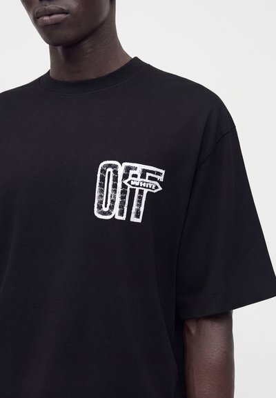 Άνδρας που φοράει μαύρο t-shirt με λευκό φθαρμένο κείμενο "OFF" και μια μικρή ετικέτα σε σχήμα βέλους με την επιγραφή "WHITE" στο αριστερό στήθος.