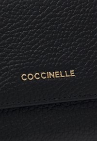 Czarna torba ze strukturalnej skóry z wytłaczanym złotym logo "Coccinelle". Gładkie wykończenie z widocznym splotem i precyzyjnym szyciem.