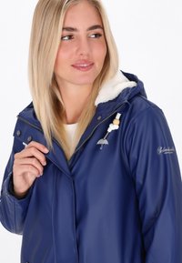 Tumši zilā ūdensnecaurlaidīgā jaka ar mākslīgās vilnas oderi, ar rāvējslēdzēju un regulējamu kapuci. Iezīmē sudraba lietussarga logotipu un dekoratīvās pērles.