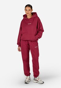 Bordový mikina s predným vreckom a zodpovedajúcimi teplákmi. Biele logo na hrudi a na boku. Textúrovaná bavlnená zmes. Model má na sebe tenisky.