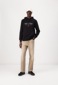 Schwarzer Hoodie mit rotem und weißem "Tommy Hilfiger"-Logo, beigefarbene gerade geschnittene Jeans und schwarze Sneakers, trägt eine hellfarbige Kappe.