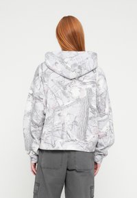 Sudadera corta con capucha en gris y blanco con un sutil patrón de ramas de árbol. Cuenta con una capucha grande y mangas extragrandes, fabricada con tela suave.