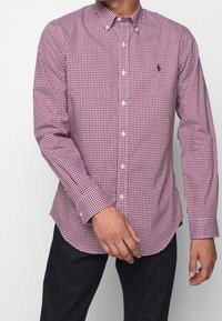 Červená a bílá kostkovaná košile na knoflíky s límečkem, dlouhými rukávy a rovnou spodní hranou. Na hrudi je výrazné malé logo.