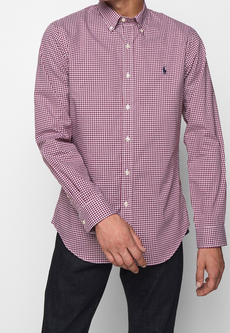 Červená a bílá kostkovaná košile na knoflíky s límečkem, dlouhými rukávy a rovnou spodní hranou. Na hrudi je výrazné malé logo.