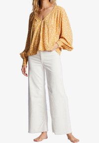 Blouse ample à motifs floraux jaunes, à manches longues et décolleté en V, associée à un pantalon en velours côtelé blanc à jambes larges.
