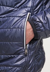 Giacca puffy blu navy con un motivo a righe orizzontali, tasca laterale con zip e dettagli della zip bianchi in contrasto. Tessuto liscio e lucido.