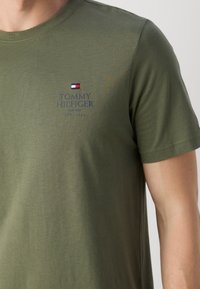 Camiseta de manga corta color verde oliva hecha de algodón, con un pequeño logotipo bordado en el pecho con texto y un detalle de bandera colorida.