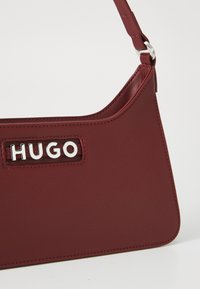Bordo kožna torba preko ramena sa srebrnim metalnim logotipom "HUGO" i tankim podesivim remenom.