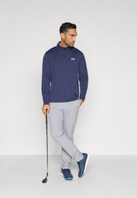 Μπλε ναυτικό πουλόβερ με φερμουάρ στο ένα τέταρτο και λευκό λογότυπο, σε συνδυασμό με ανοιχτό γκρι παντελόνι και σκούρα μπλε παπούτσια γκολφ, κρατώντας ένα μπαστούνι γκολφ.