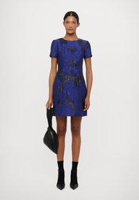 Zila rakstainā kleita ar melniem akcentiem, īsām piedurknēm un pieguļošu siluetu. Valkāta ar melnām potīšu zeķēm un norādītām kurpēm, turētā melna soma.