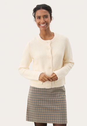 Vaaleanbeige neulecardigan, jossa on tekstuurinen kuvio, pyöreä pääntie ja viisi nappia. Yhdistetty kuvioituun ruutuhameeseen.