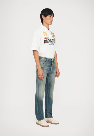 T-shirt bianca in cotone con stampe grafiche e scritte blu, abbinata a jeans in denim blu scoloriti e sneakers di colore chiaro.