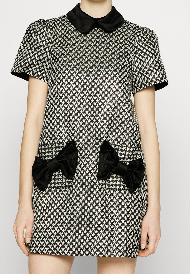 Kortärmad klänning i svart och silver houndstooth-mönster med svart satinkrage och stora svarta rosetter på framfickorna.