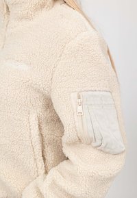 Pullover in pile beige con una superficie morbida e текстurizzata; presenta una tasca con cerniera sulla parte superiore del braccio e un logo discreto sul petto.