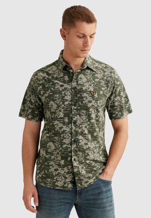 Homme portant une chemise verte à manches courtes à boutons avec un motif floral abstrait et un emblème brodé d'oiseau jaune et noir, associée à un jean bleu.