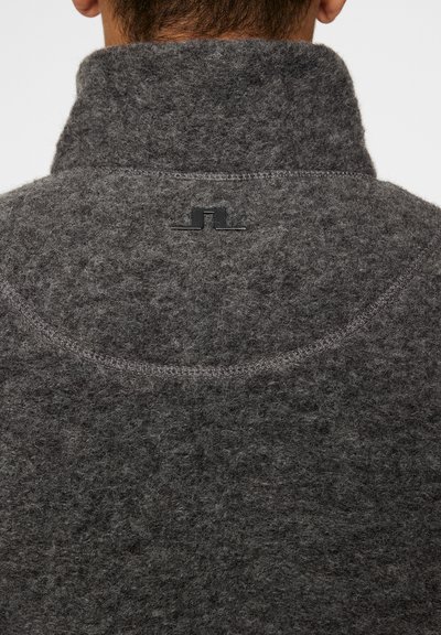 Grå uldpullover med høj krave, der har en struktureret, fuzzy overflade og et lille sort logo bagpå kraven. Synlige stikninger.