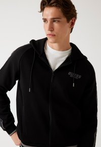 Felpa nera con zip realizzata in tessuto morbido, con cerniera frontale, cappuccio con coulisse e dettagli in grigio sul logo al petto e sulle strisce delle maniche.