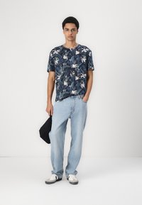 Tricou bleumarin cu mânecă scurtă, decorat cu un model floral tropical, asortat cu blugi albaștri deschis și o geantă neagră. Tenisi în culori alb și gri.