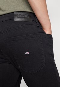 Persona vistiendo jeans negros de Tommy con la mano en el bolsillo, visible etiqueta de la marca en la cinturilla y logotipo bordado en el bolsillo trasero.