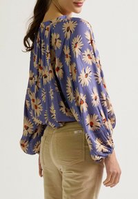 Lila Bluse mit floralem Muster, die große cremefarbene Gänseblümchen mit roten Zentren zeigt. Mit aufgekrempelten Ärmeln und lockerer Passform. Kombiniert mit beigen Cordhosen.