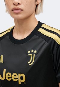 Personne portant un maillot de football noir de la Juventus avec des bandes dorées sur les épaules, un logo Jeep doré et un emblème de l'équipe sur la poitrine.