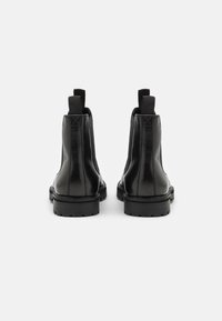 Botas de tobillo de cuero negro con paneles laterales elásticos y tiradores, vistas desde atrás sobre un fondo blanco.