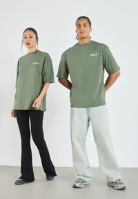 Oliven grønne oversized T-shirts har et lille "PARIS" logo. Kvinden har sorte ribbede flared bukser på, mens manden har lyseblå jeans og grå sneakers.